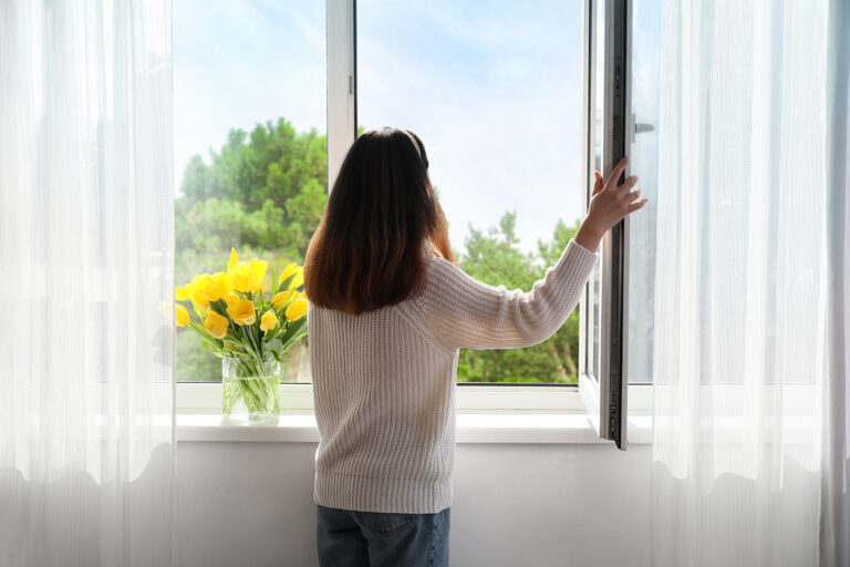 Pretty young Asian woman opening window at home on sunny day, ba