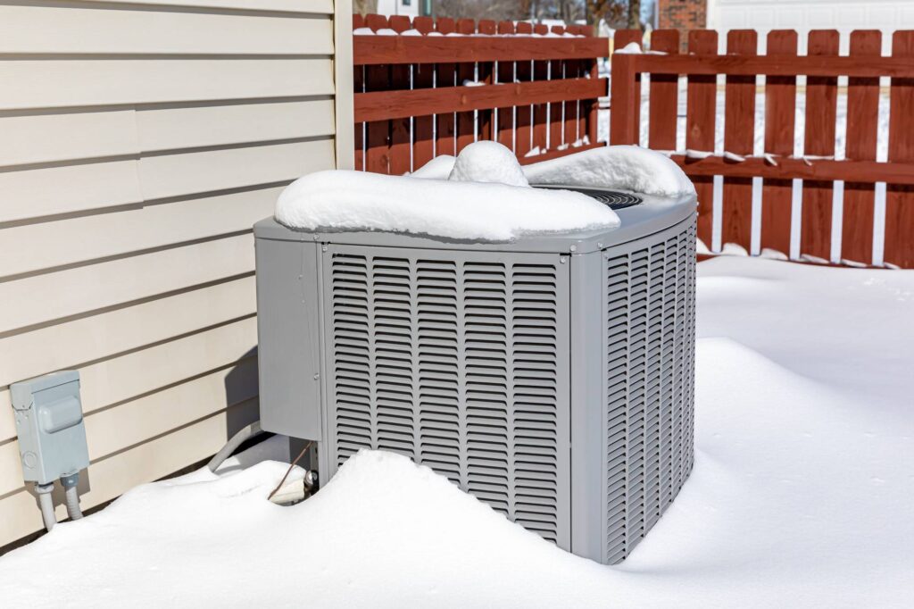 Outdoor air conditioning unit covered in snow during winter weather beside a house