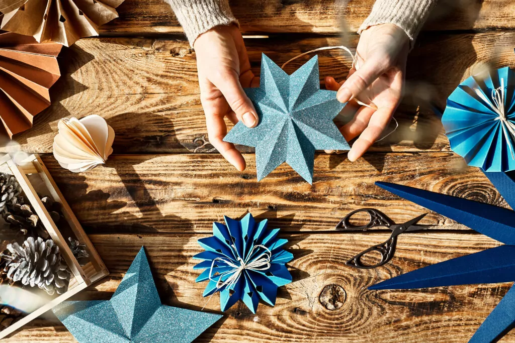 Winter holiday decoration. Woman's hands making christmas paper snowflakes, paper blue stars and fans made of craft paper on wooden table. Nature aesthetic. Zero waste handmade gifts. DIY.