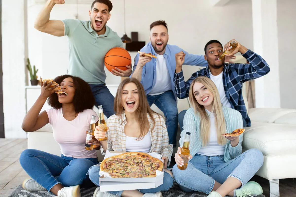 Group of diverse young friends with pizza and beer watching basketball game on TV, supporting favorite team at home